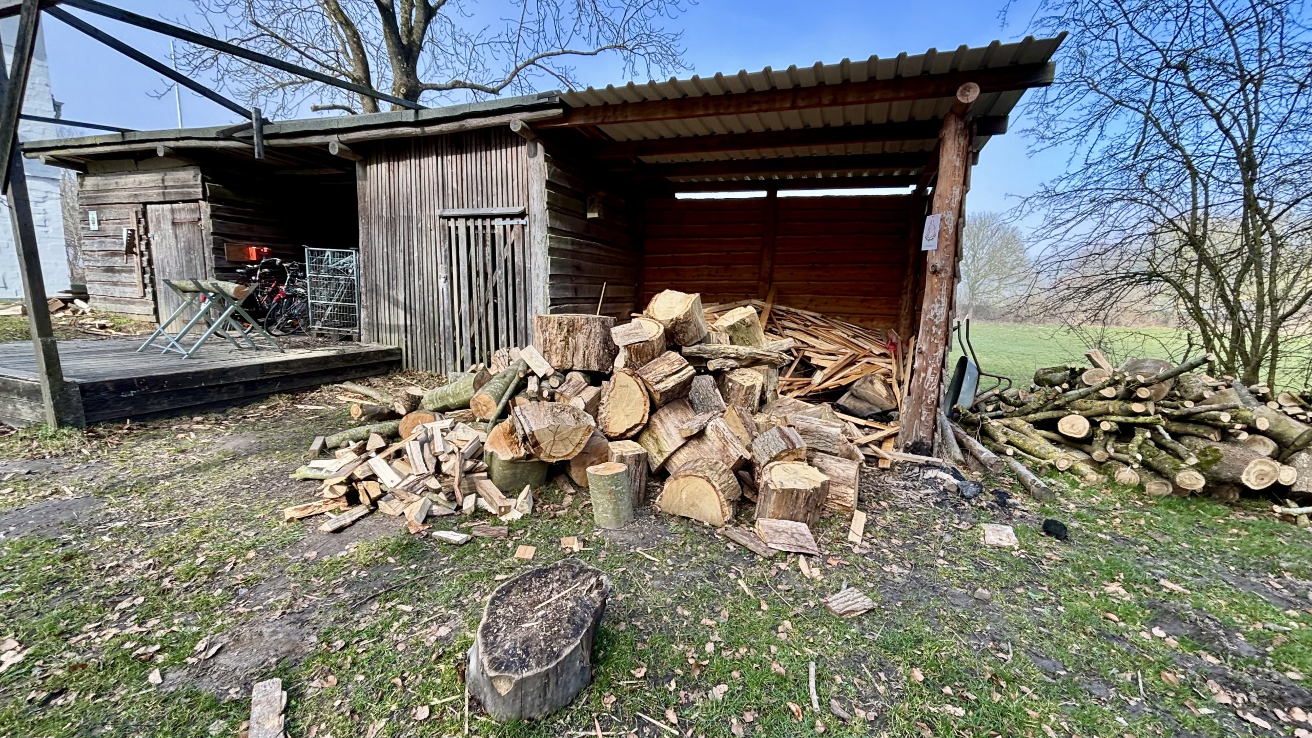 Foto eines Holzschuppens auf einer grünen Wiese. Vor dem Schuppen liegt ein Haufen Holz, teilweise schon zu Brennholz gespalten. Links ein Bock mit einem großen Holzstück, dahinter Fahrräder. Im Bildvordergrund steht ein Hackklotz.